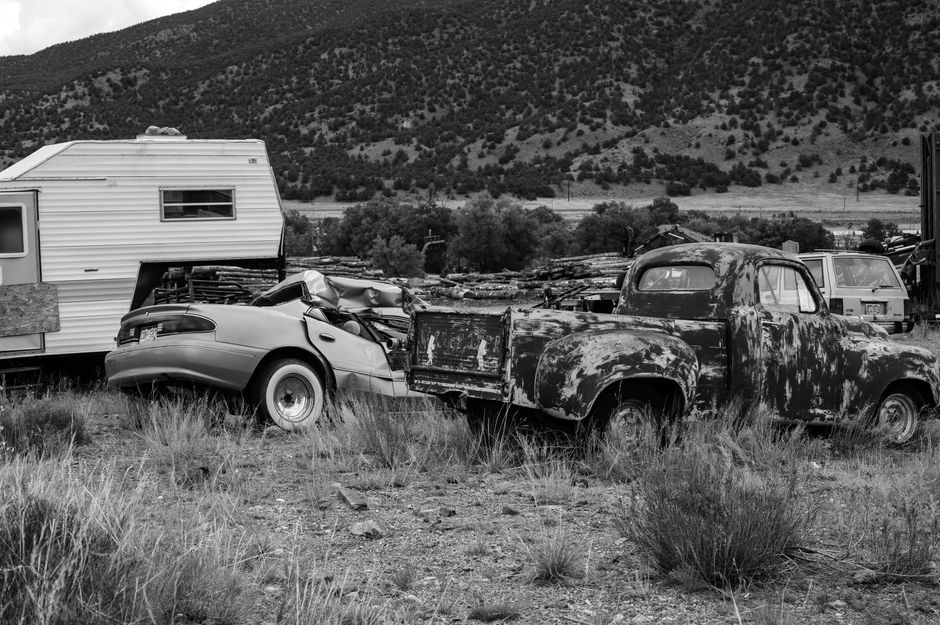 Black and white photo of rusty abandoned vehicles and camper in rural setting.