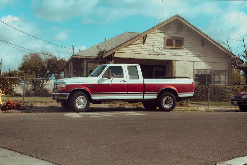Classic red and white Ford pickup truck parked on an empty street in front of a rustic house.